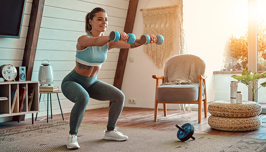 A woman lifting dumbbells indoors, focusing on fitness, with a chair and blanket visible in the background.
