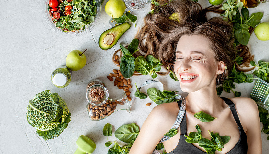 A woman smiling, surrounded by fruits and vegetables, lies on the floor, embodying a healthy lifestyle aligned with SteelFit USA's fitness goals.