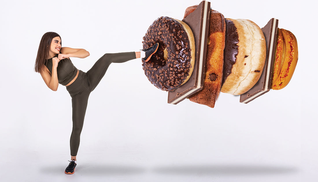 A woman playfully kicks a large chocolate-covered donut, highlighting an energetic and dynamic approach to fitness and indulgence.