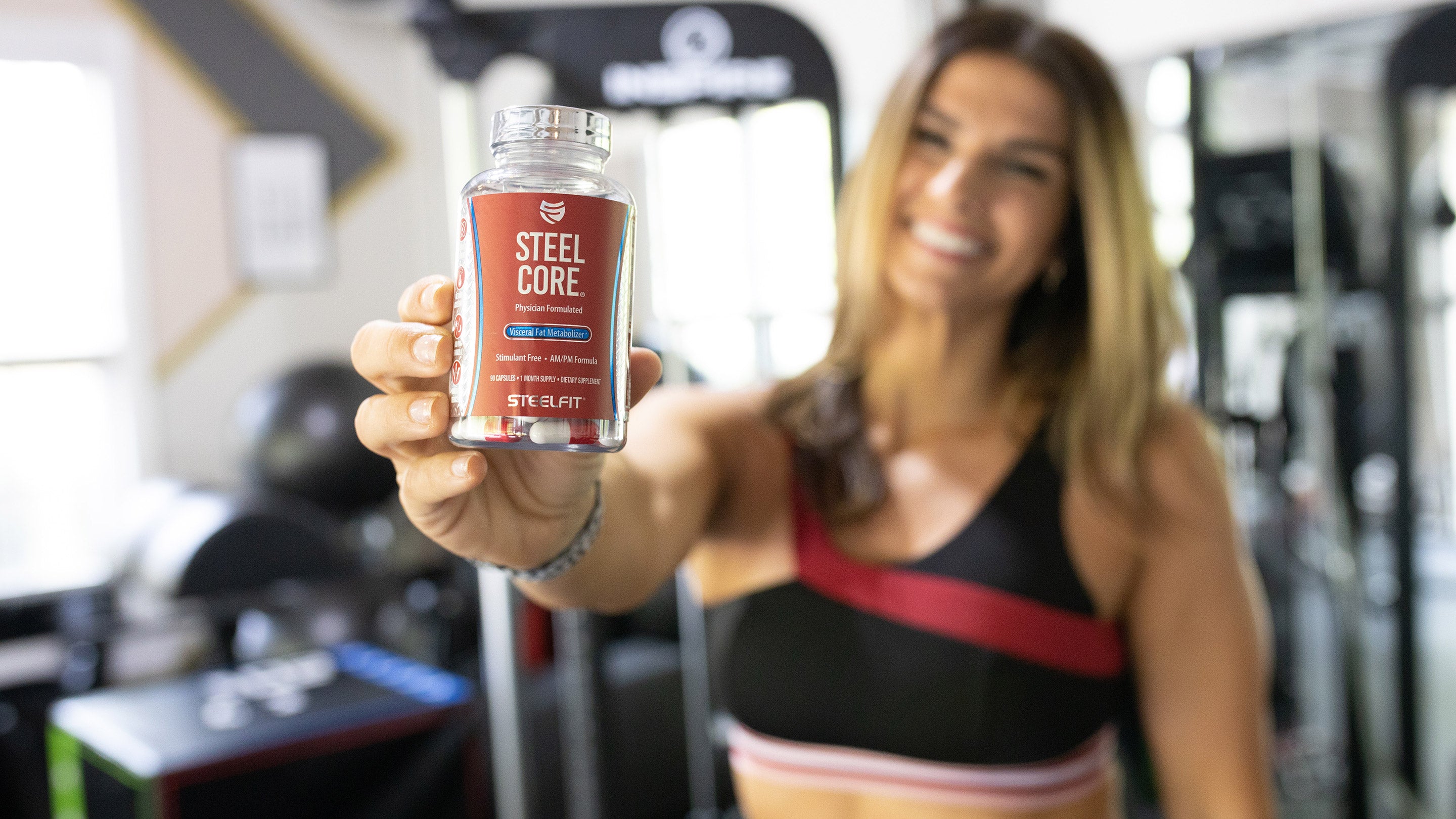 A woman smiling while holding a bottle of Steel Core, a sports nutrition supplement from SteelFit USA.