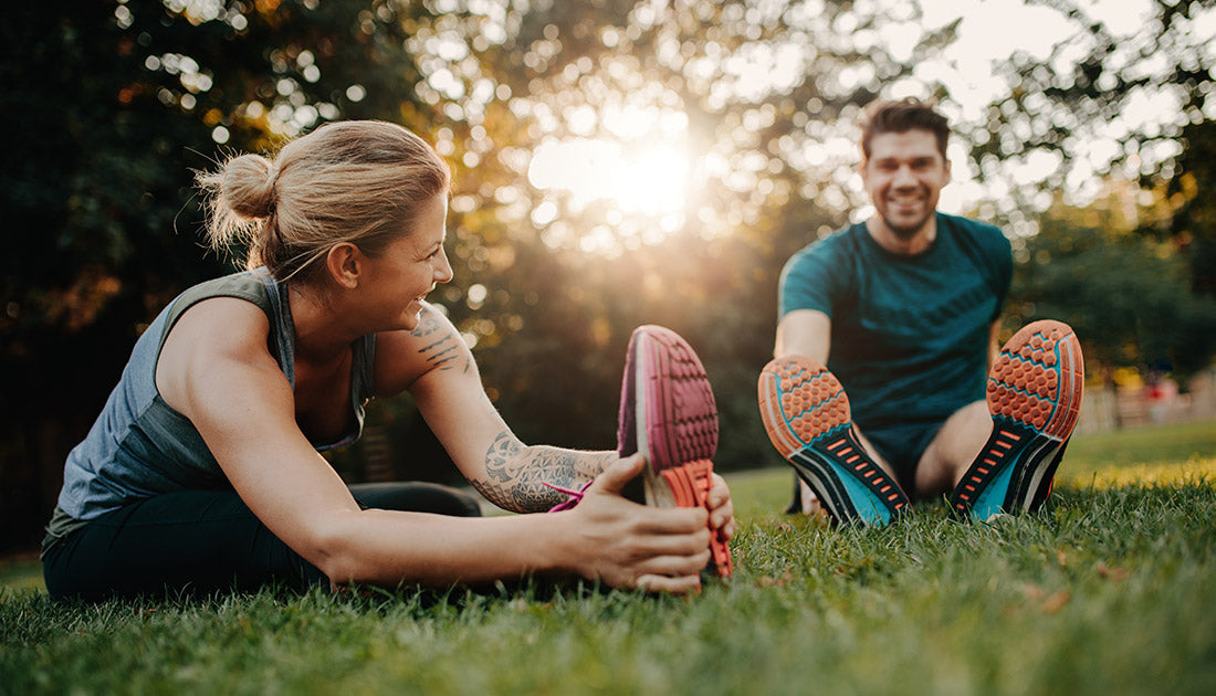 A man and woman stretching on grass, wearing sporty attire, with a focus on vibrant footwear, reflecting an active, health-focused lifestyle.