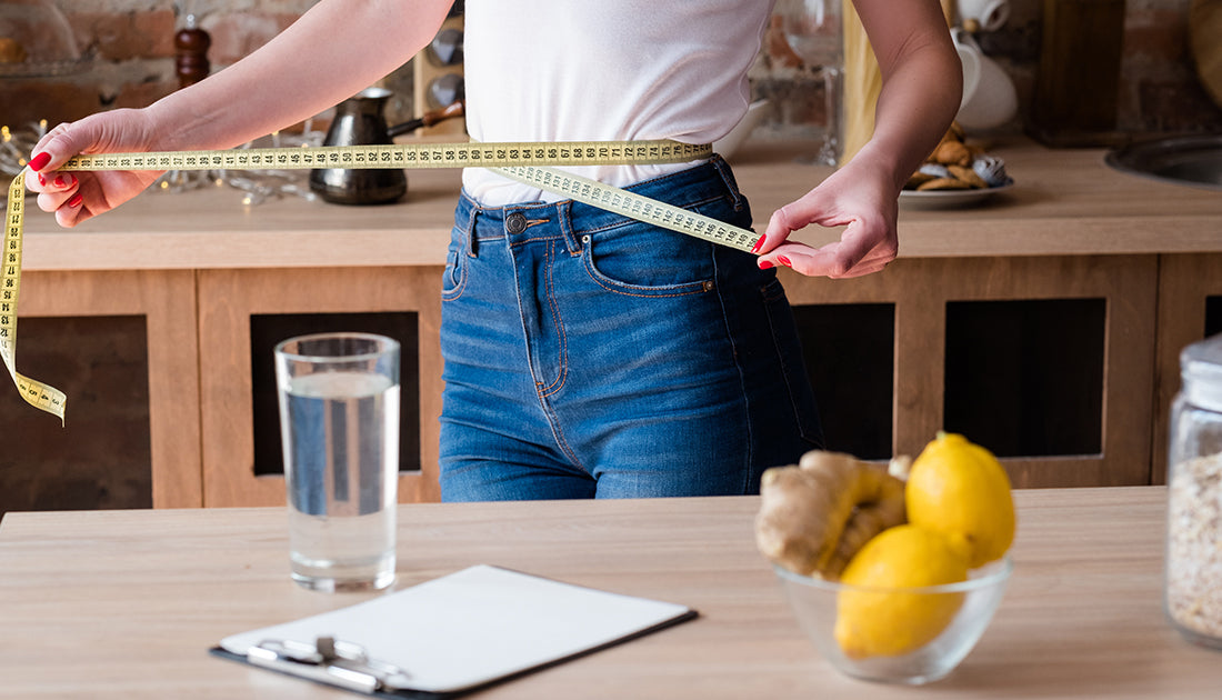 A person measures their waist, with lemons and ginger in a bowl nearby, reflecting a focus on health and fitness.