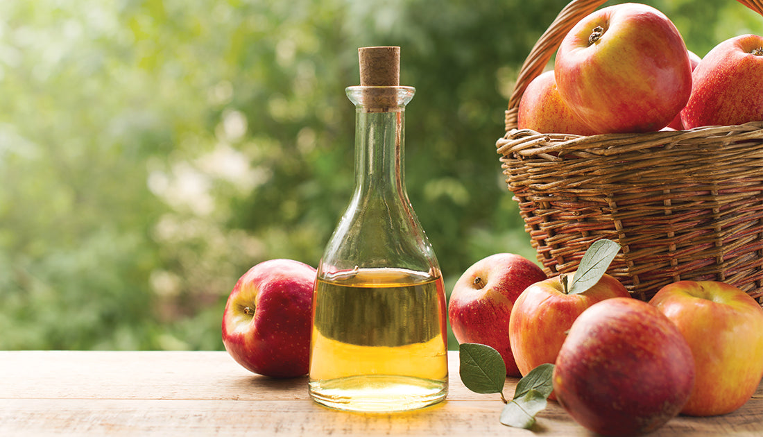 A glass bottle of apple cider vinegar next to a basket of fresh apples