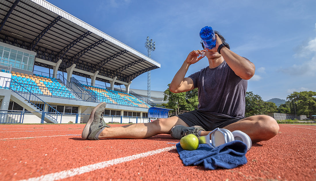 A man on a track drinks from a blue bottle, with a towel and green apple beside him, suggesting a fitness-focused lifestyle.