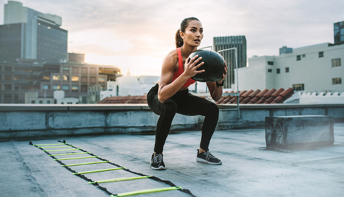 Female athlete doing squats holding a medicine ball standing on a rooftop. Woman doing workout using medicine ball with an agility ladder by her side on rooftop.