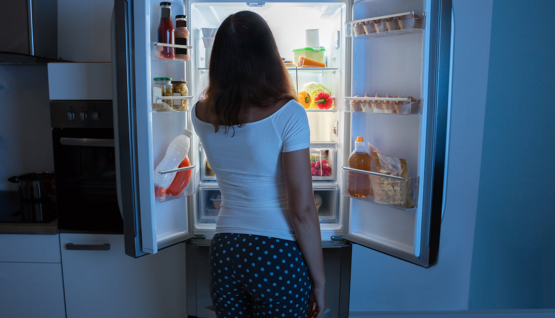 A woman stands in front of an open refrigerator, examining contents, reflecting a focus on health and fitness through nutrition.