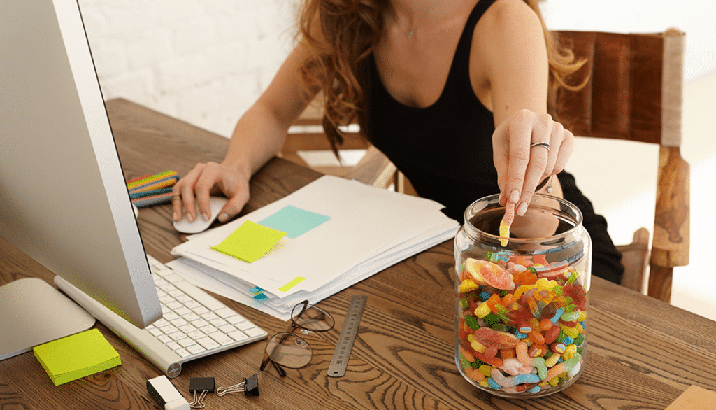 A woman sits at a table with a jar of candy and papers, suggesting a casual workspace or break moment.