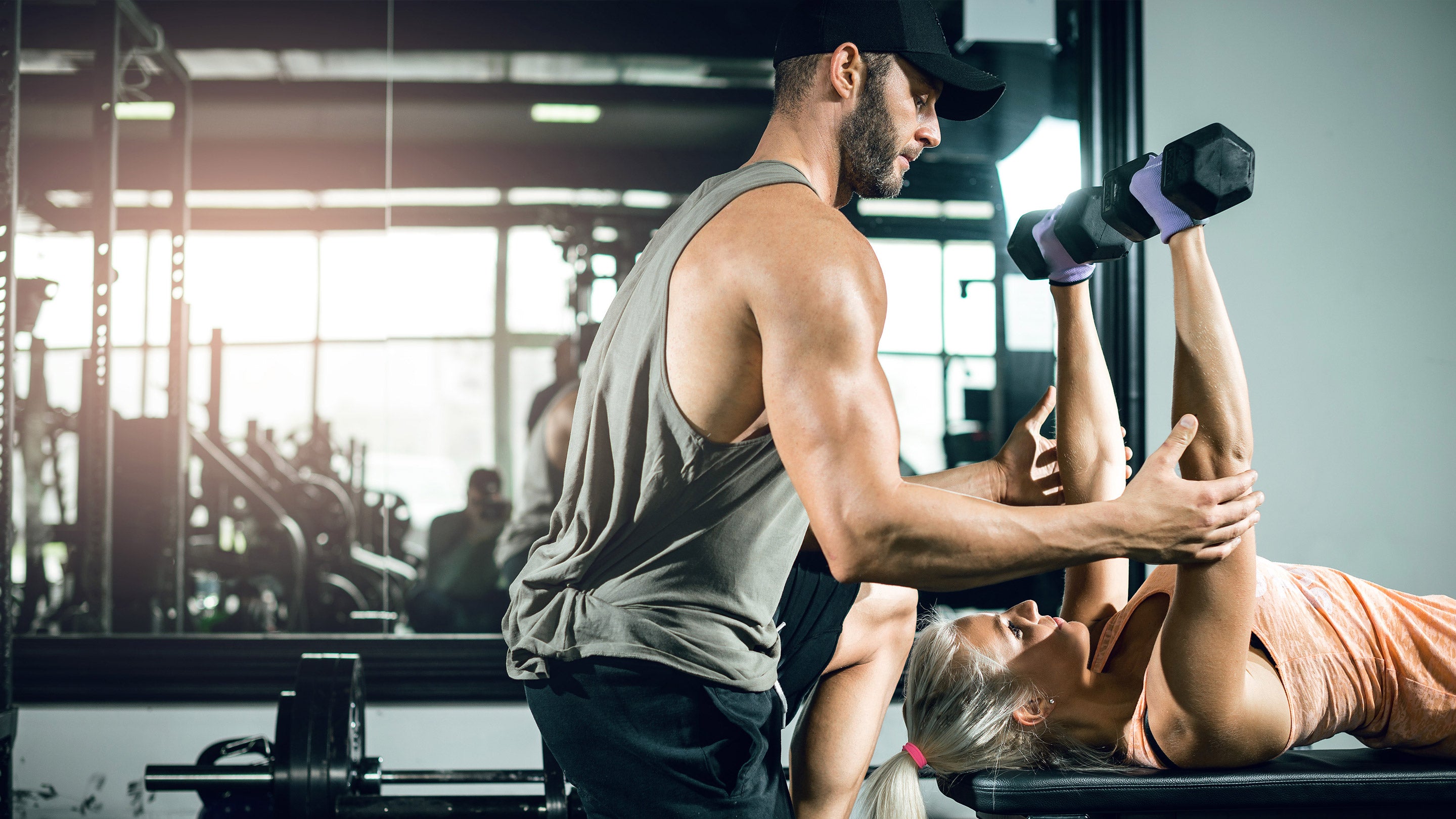 A man assists a woman with weightlifting in a gym setting, reflecting a focus on physical fitness and strength training.