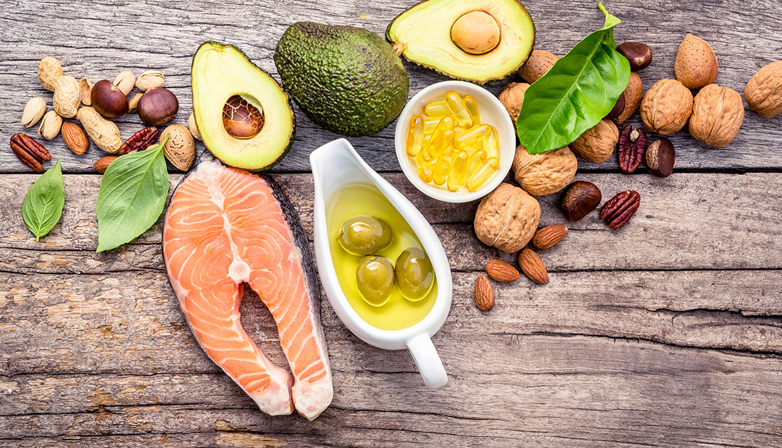 A variety of food ingredients, including salmon, avocado, olives, and a bowl of yellow pills, arranged on a table.