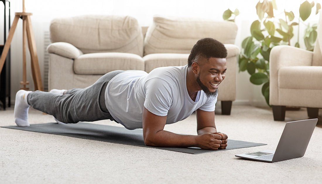 A man doing push-ups on a mat near a couch, with a laptop on the floor, in a fitness-focused indoor setting.