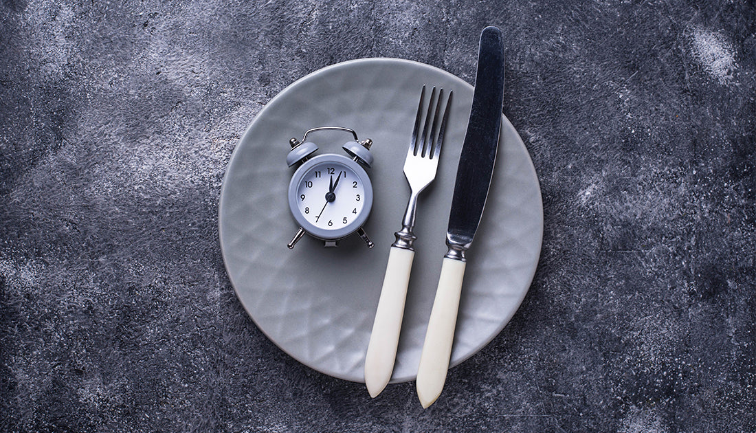 A white alarm clock on a plate with a knife and fork, symbolizing time management in fitness and nutrition.