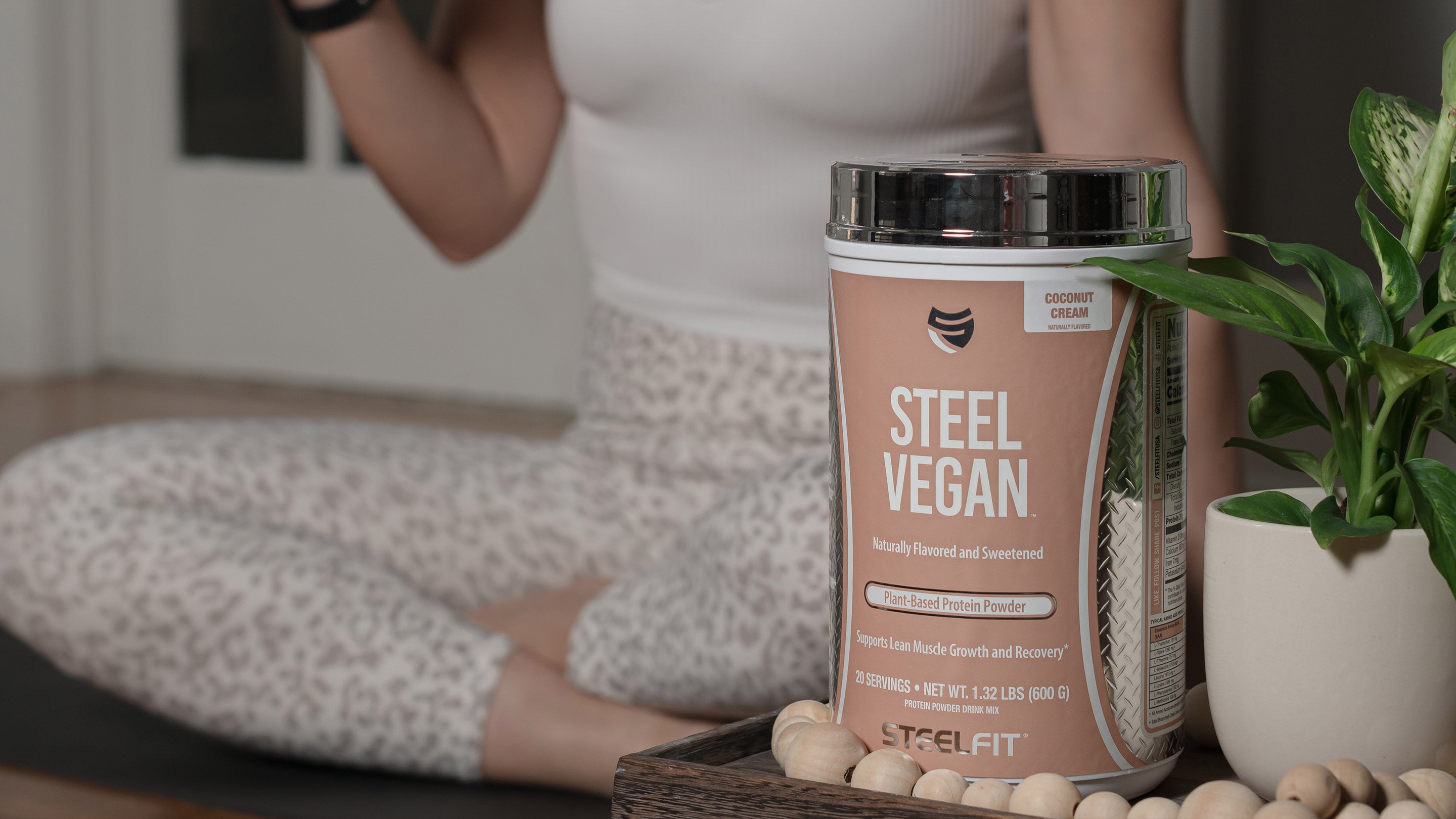 A woman sits on a bed holding a container of coconut cream, with a plant in the background, reflecting a fitness and wellness theme.