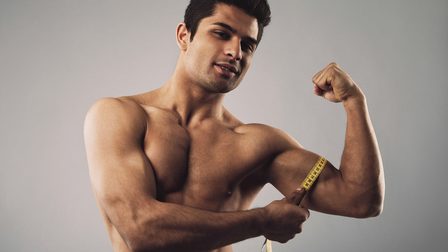 Fit hispanic male model measuring his body. Masculine young male measuring biceps with tape measure on grey background.