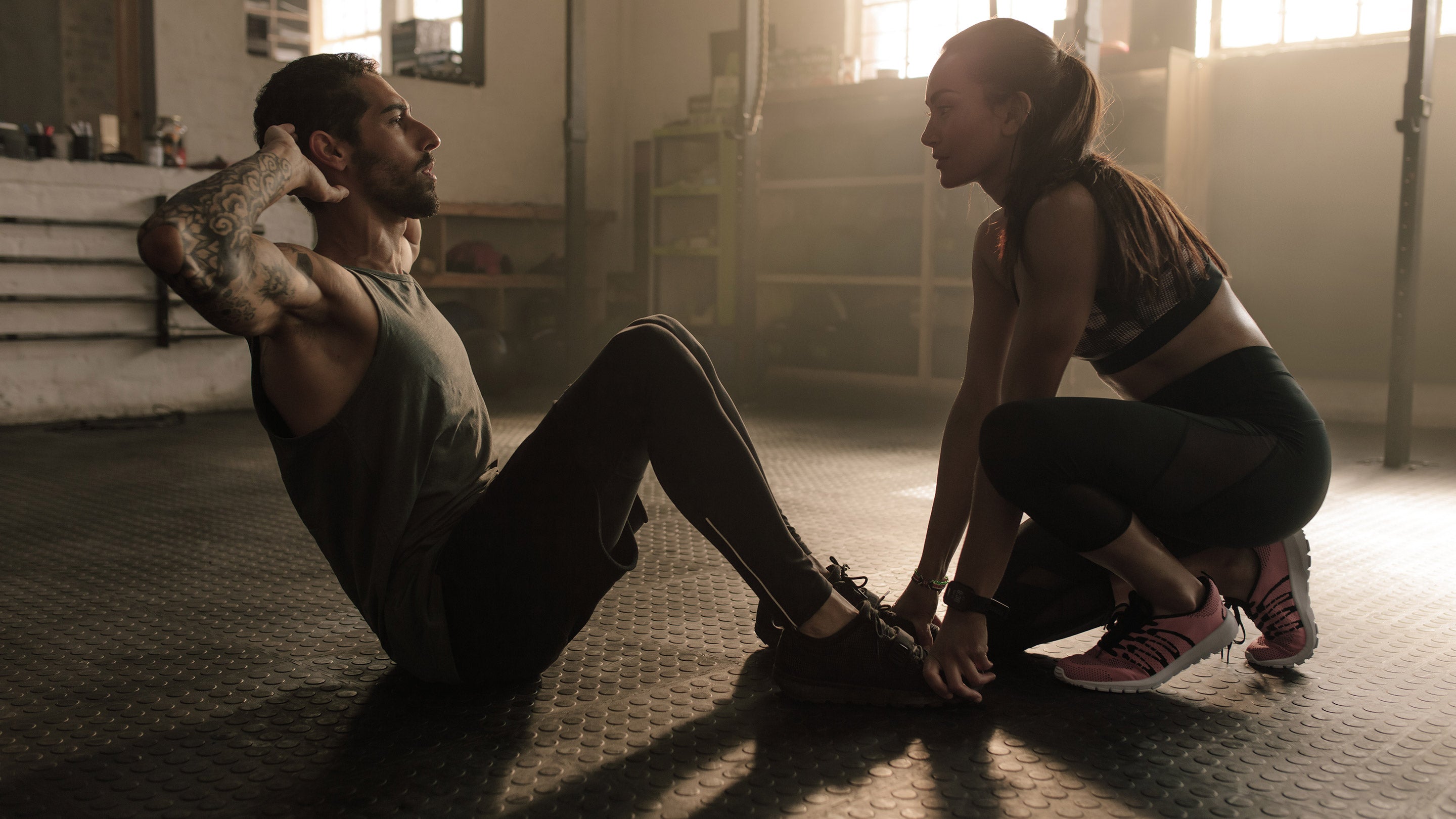 A man and woman sitting on the floor; the woman is tying her pink and black shoes, suggesting a fitness setting.