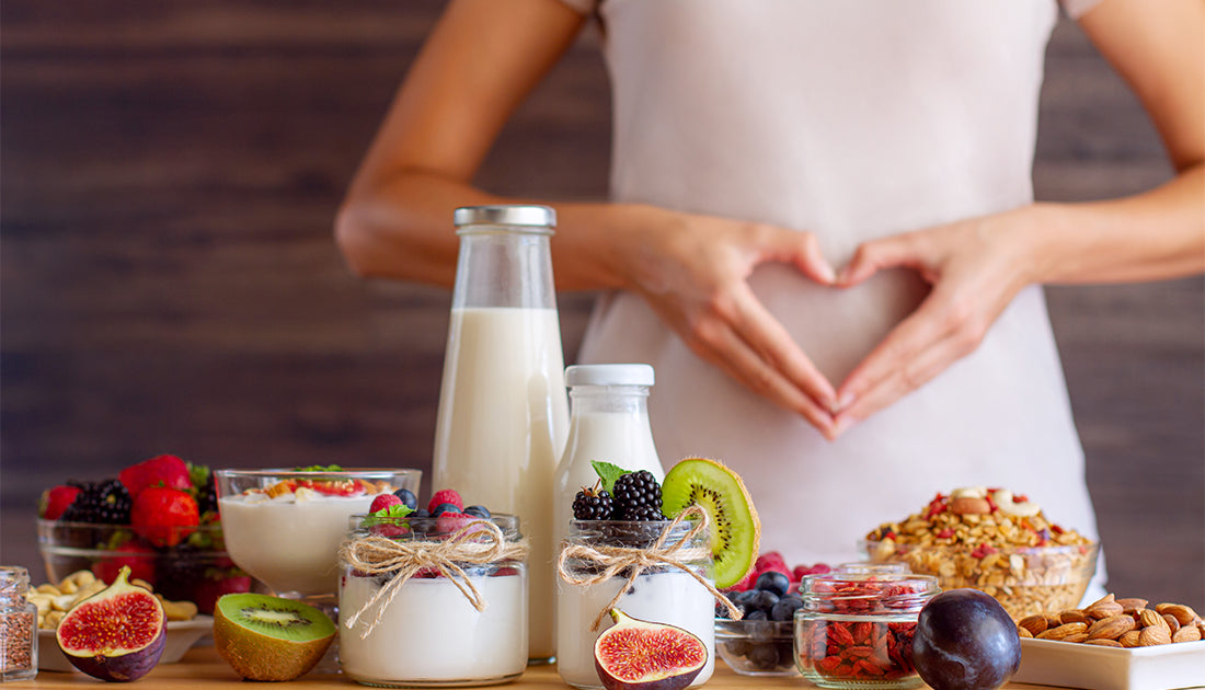 A woman forms a heart with her hands over a table of nutritious foods including milk, goji berries, and assorted fruits.