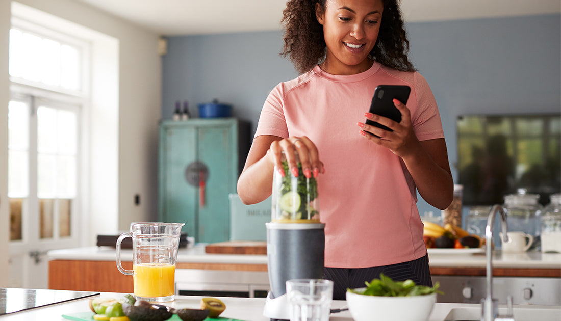 A woman blends a smoothie while using her phone, embodying a healthy lifestyle focus, relevant to fitness and nutrition.