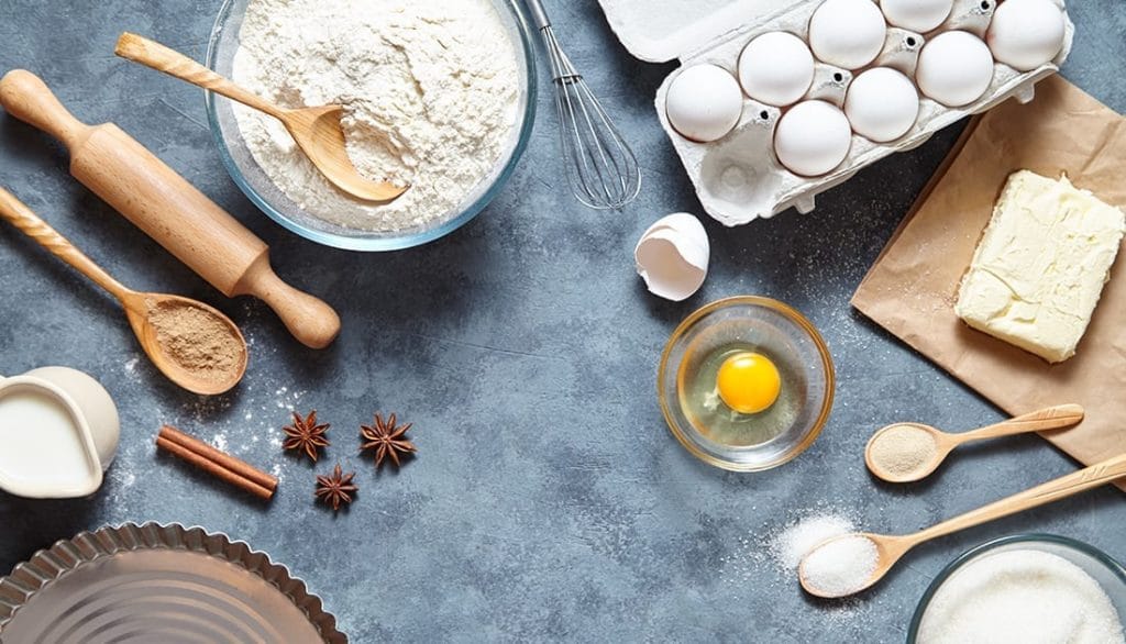 A table with flour, a wooden spoon, rolling pin, egg yolk, and a white pitcher, representing ingredients for fitness-focused meal preparation.