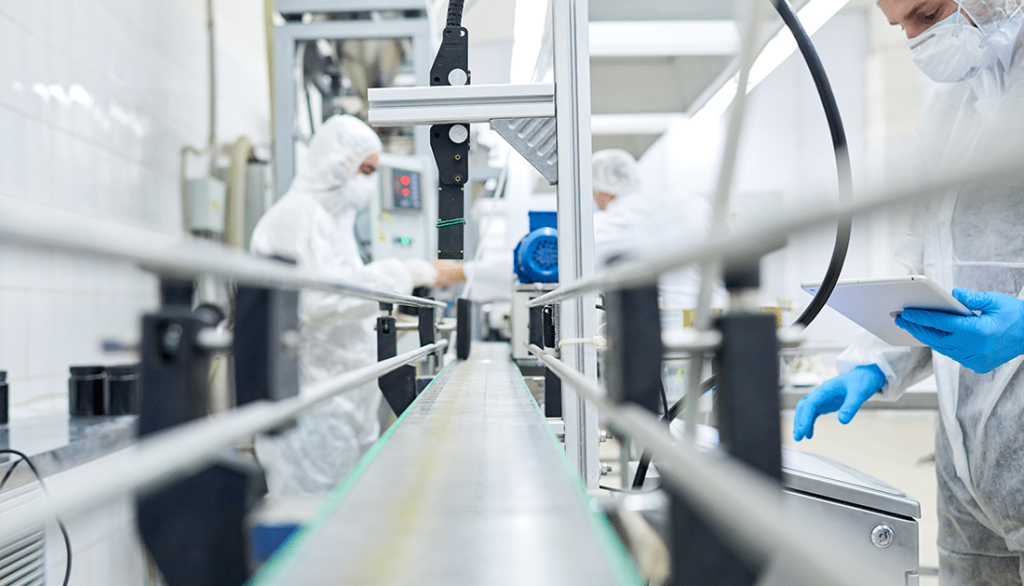 A technician in a white hazmat suit and blue gloves works on a conveyor belt, holding a tablet in a laboratory setting.