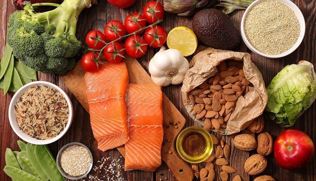 An assortment of healthy foods on a table, including a bowl of quinoa, oatmeal, broccoli, salmon, a red apple, and a lemon.