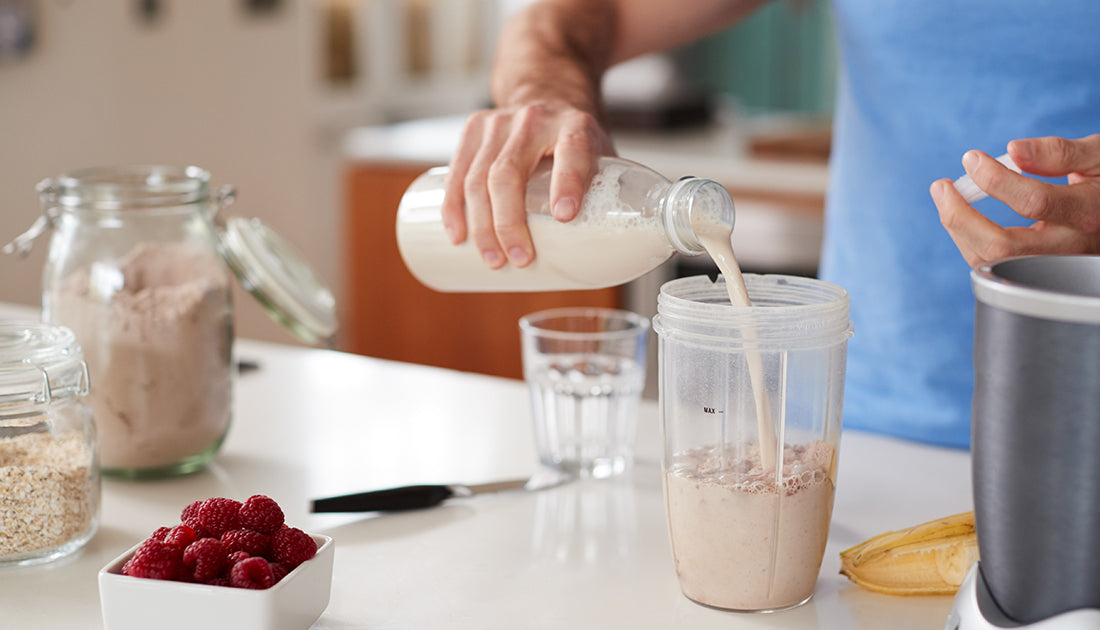 A person pours milk into a cup near a blender, with a bowl of raspberries on the table, reflecting a health-focused kitchen setting.