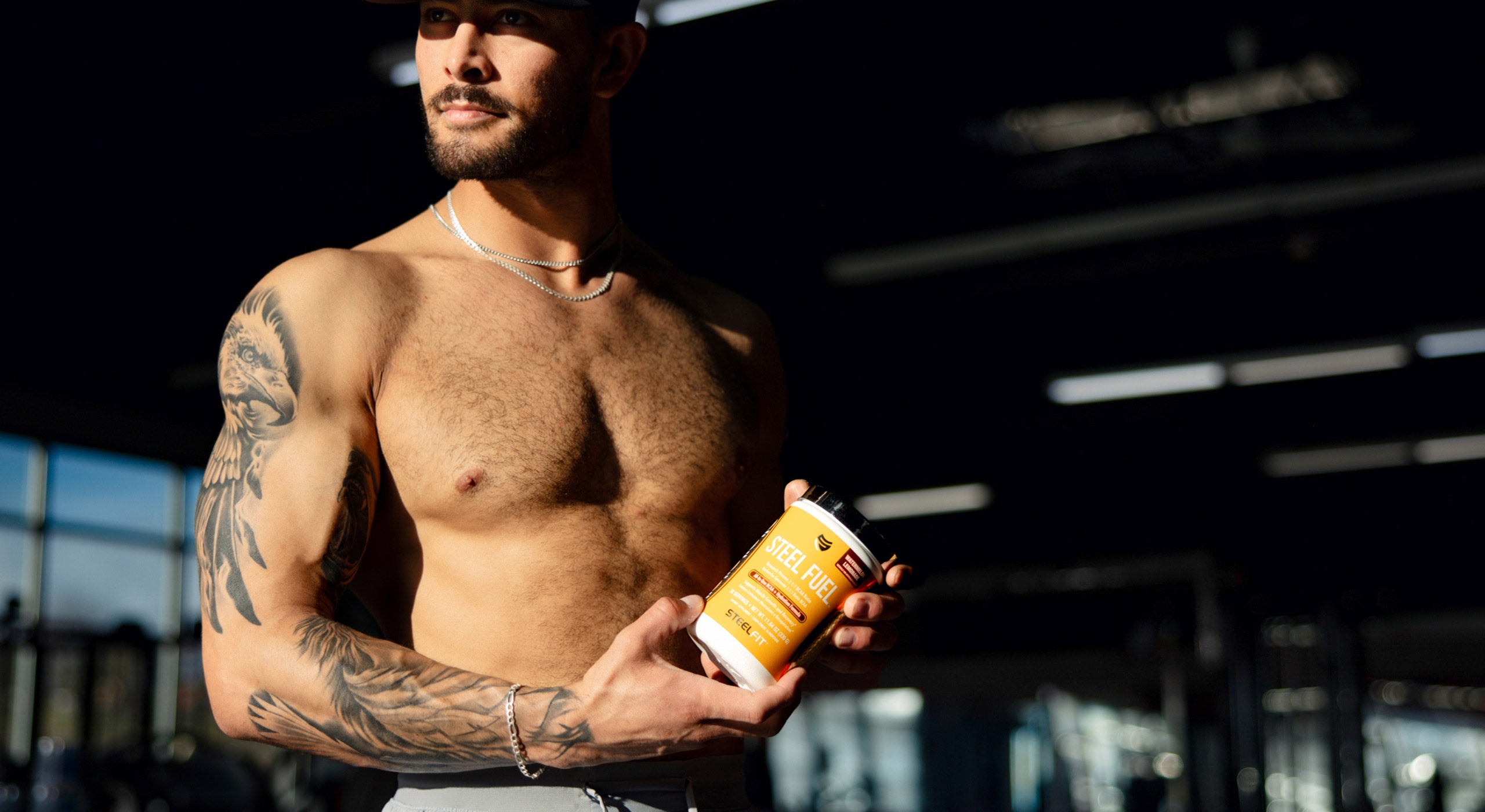 A shirtless man with tattoos holds a bottle of protein, reflecting sports nutrition focus.