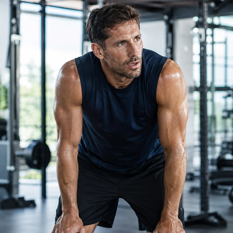 A muscular man in a gym wearing a dark navy sleeveless athletic shirt and black shorts, leaning forward with a focused expression.